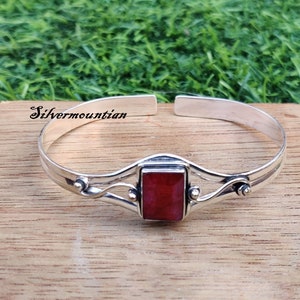 May include: Silver bracelet with a red stone set in a silver frame. The bracelet has a delicate design with two curved lines and small silver beads.