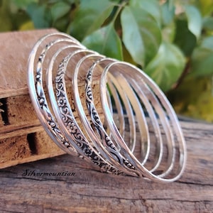 May include: A set of seven silver bangles with different decorative patterns. The bangles are stacked on top of each other and have a shiny finish.