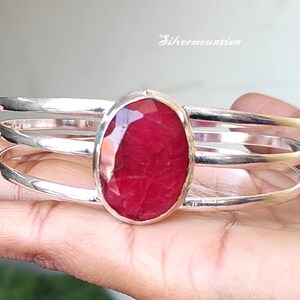 May include: A silver bangle bracelet with a large, oval-shaped, red gemstone set in the center. The bracelet has three bands of silver.