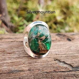 May include: A silver ring with a large, oval-shaped, green and brown stone. The stone has a textured appearance. The ring is set on a wooden surface. The text "Silvermountian" is visible in the image.