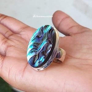 May include: A silver ring featuring an oval abalone shell inlay. The shell displays a blue, green, and black iridescent pattern.