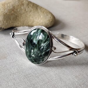 Könnte beinhalten: Ein silbernes Armreif-Armband mit einem ovalen, grün-weißen Edelstein. Der Edelstein hat ein wirbelndes Muster.