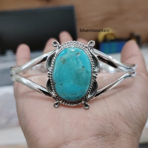 Può includere: Un bracciale a polsino in argento con una grande pietra ovale turchese al centro. Il turchese è di un vivace colore blu-verde, incastonato in una cornice d'argento decorativa. Il bracciale ha un design semplice ed elegante.