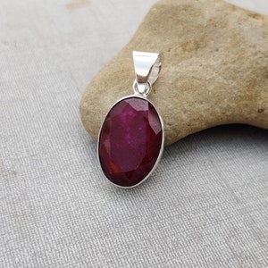 May include: A silver pendant featuring a large, oval-shaped ruby gemstone. The ruby is a deep, rich red color and has a faceted cut.