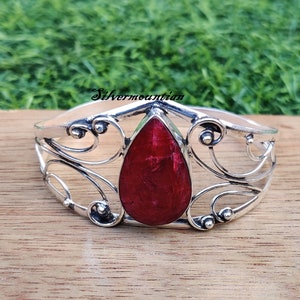 Könnte beinhalten: Ein silbernes Armreif-Armband mit einem roten Edelstein in der Mitte. Das Armband hat ein zartes, verziertes Design.