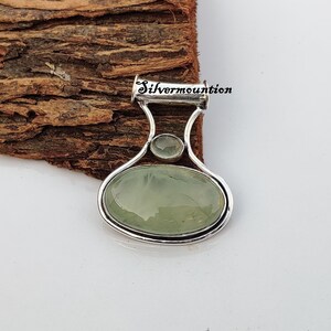 May include: A silver pendant featuring a green oval gemstone and a smaller round green gemstone. The pendant is displayed on a wooden surface. The text "Silvermounton" is visible on the pendant.