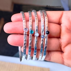 Pode incluir: Cinco pulseiras de prata martelada com pedras preciosas de diferentes cores. As pedras preciosas são ametista, verde, branco, pedra da lua e preto.