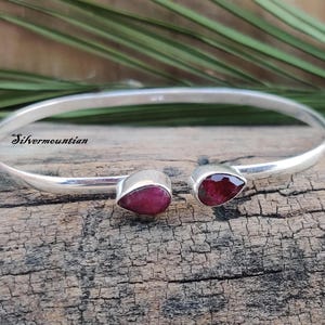 May include: A silver bangle bracelet with two teardrop-shaped red gemstones. The bracelet is open and has a simple design.