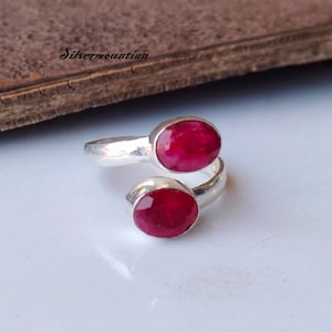 May include: A silver ring with two oval, vivid red gemstones. The gemstones are set in a wrap-around design, giving a modern and elegant appearance. The ring is displayed on a white surface.