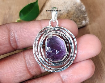 May include: A silver pendant with a purple amethyst gemstone set in a circular design with concentric circles. The pendant is on a hand.