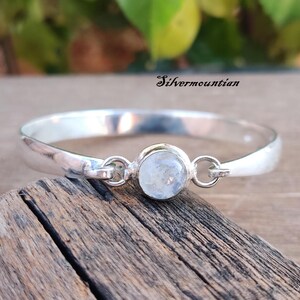 May include: A silver bangle bracelet with a round moonstone charm. The bracelet is open and has a hinged clasp.