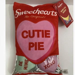 May include: A red Sweethearts candy heart pillow with the words "CUTIE PIE" on a pink heart. The pillow also features two smaller heart-shaped candies that say "BE MINE" and "XOXO". A tag reads "MORE FUN INSIDE!"