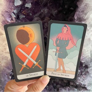 May include: Two tarot cards are displayed, featuring illustrations and text. One card shows crossed swords over a heart with a crescent moon. The other depicts a figure holding a sword. The cards are set against a purple background.
