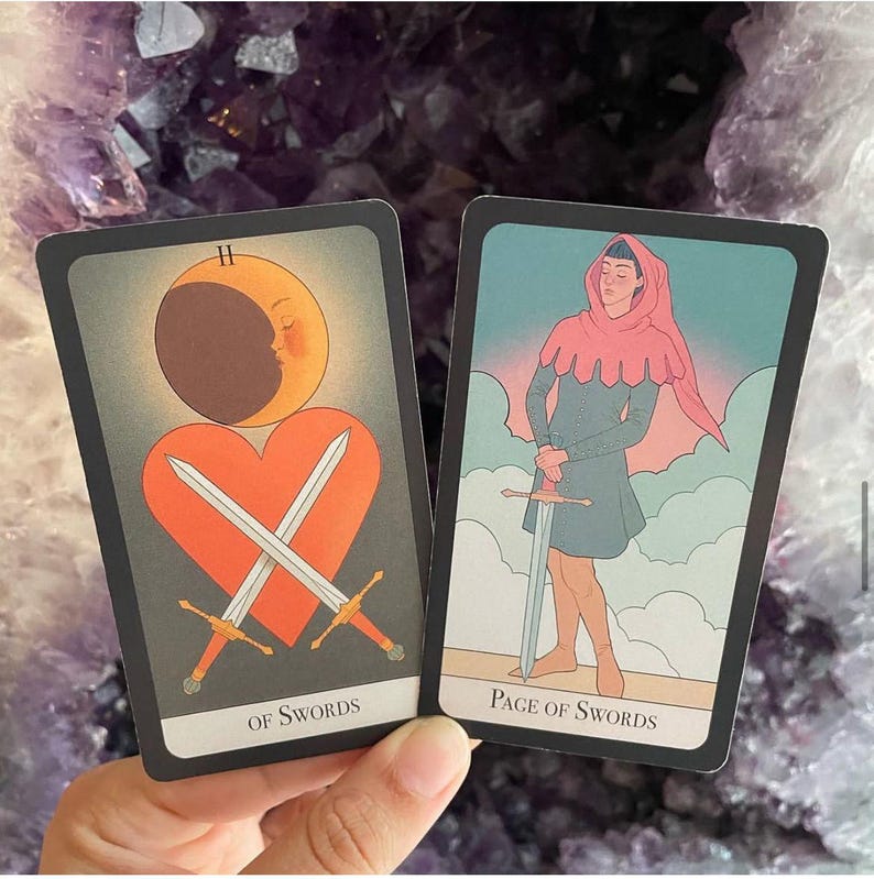 May include: Two tarot cards are displayed, featuring illustrations and text. One card shows crossed swords over a heart with a crescent moon. The other depicts a figure holding a sword. The cards are set against a purple background.