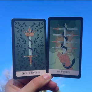 May include: Two tarot cards against a blue sky. The "Ace of Swords" card features a sword with a crown and snake, surrounded by leaves. The "Of Swords" card shows a hand holding a sword with other swords in the background. Both cards have text.