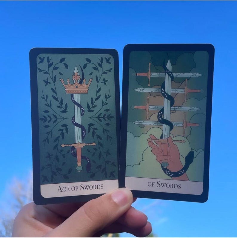 May include: Two tarot cards against a blue sky. The "Ace of Swords" card features a sword with a crown and snake, surrounded by leaves. The "Of Swords" card shows a hand holding a sword with other swords in the background. Both cards have text.