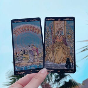 May include: Two tarot cards are held in a hand. The cards have black borders and feature detailed illustrations. One card depicts a couple under an archway, and the other shows a queen with a black cat. Both cards have the number 10.