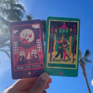 May include: Two tarot cards are held up against a blue sky. The card on the left is purple with a moon and cat illustration, and the text "XVIII LA LUNE". The card on the right is green with a couple dancing and the text "Four of Wands".