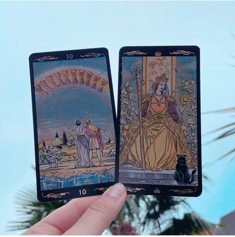 May include: Two tarot cards are held in a hand. The cards have black borders and feature detailed illustrations. One card depicts a couple under an archway, and the other shows a queen with a black cat. Both cards have the number 10.