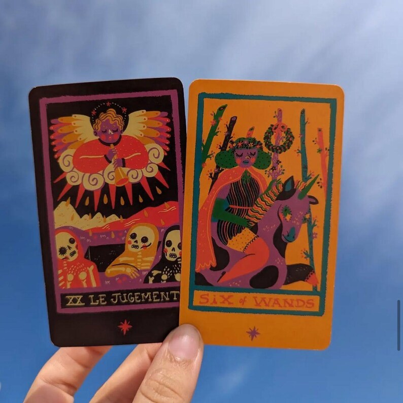 May include: Two tarot cards are displayed against a blue sky. One card is black with a purple border and features an angel and skeletons. The other card is orange with a green border and depicts a person riding a purple unicorn. The text "XX LE JUGEMENT" and "SIX of WANDS" are visible.