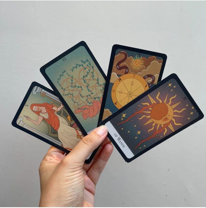 May include: A hand holds a fan of tarot cards with various illustrations. The cards feature images of a wheel, a sun, and other symbolic designs in shades of blue, orange, and beige. The card labeled "Of Wands" is visible.