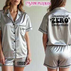 May include: Silver satin pyjama set with black trim. The short-sleeved top has a breast pocket with the name "Caitlyn" embroidered. The shorts have the words "CHEER LEADER" printed. The back of the top says "Dreaming of, ZERO Deductions".
