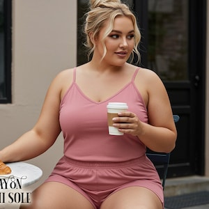 Può includere: Un completo camisole e pantaloncini rosa cipria. La camisole ha uno scollo a V e spalline sottili. I pantaloncini sono in tinta. Si tiene in mano una tazza di caffè di carta bianca. Un croissant è su un tavolino bianco. Il testo "RAYON CAMI SOLE" è in basso.