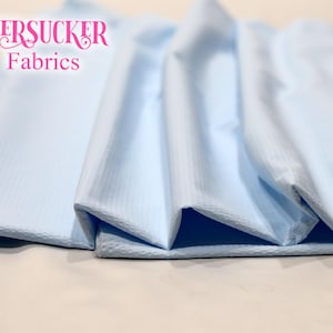 Puede incluir: Tejido seersucker azul claro, doblado y expuesto. El tejido tiene un patr&oacute;n texturizado y a rayas. Las palabras "SEERSUCKER Fabrics" se muestran en rosa, en fuente cursiva en la parte superior izquierda de la imagen.