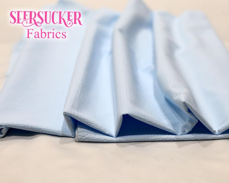 Puede incluir: Tejido seersucker azul claro, doblado y expuesto. El tejido tiene un patr&oacute;n texturizado y a rayas. Las palabras "SEERSUCKER Fabrics" se muestran en rosa, en fuente cursiva en la parte superior izquierda de la imagen.