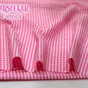 Puede incluir: Primer plano de tela seersucker rosa y blanca con estampado de cuadros. La tela est&aacute; drapeada, mostrando pliegues y textura. Las palabras "SEERSUCKER Fabrics" est&aacute;n impresas en una fuente de escritura en la parte superior.