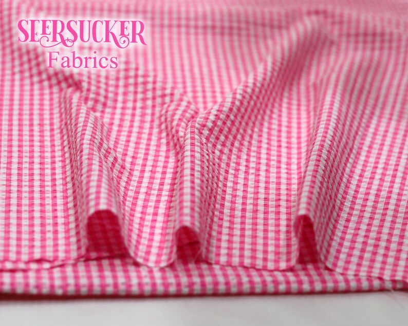 Puede incluir: Primer plano de tela seersucker rosa y blanca con estampado de cuadros. La tela est&aacute; drapeada, mostrando pliegues y textura. Las palabras "SEERSUCKER Fabrics" est&aacute;n impresas en una fuente de escritura en la parte superior.