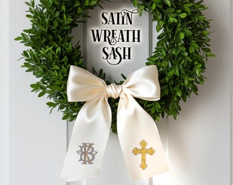 Könnte beinhalten: Ein grüner Kranz mit einer cremefarbenen Satinschleife und Schärpe. Auf der Schärpe steht der Text "SATIN WREATH SASH" in Schwarz. Ein Schärpenende hat ein goldenes Kreuz, das andere gestickte Initialen. Ein dekoratives Element für Türen oder Wände.