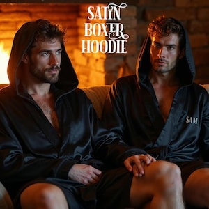 May include: Two people wearing black satin boxer hoodies with the word "SAM" embroidered on the left chest. The hoodies have attached hoods and are worn open. The image is set in front of a fireplace.