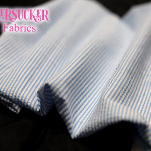 Puede incluir: Primer plano de tela seersucker con rayas azules y blancas. La tela est&aacute; doblada, mostrando la textura y el patr&oacute;n. El texto "SEERSUCKER Fabrics" est&aacute; en rosa en cursiva.
