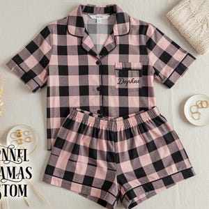 May include: Pink and black plaid flannel pajama set. The short-sleeved top has a breast pocket with the name "Daphne" embroidered on it. The matching shorts have an elastic waistband. The words "FLANNEL PAJAMAS CUSTOM" are printed on the left side.
