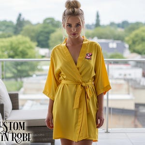 May include: A yellow satin robe with a matching belt. The robe features a small embroidered design on the front. The text "CUSTOM SATIN ROBE" is displayed in a stylized font on the lower left corner of the image.