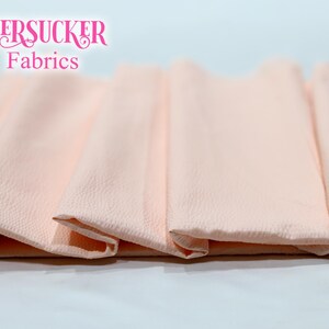 Puede incluir: Primer plano de tela seersucker melocot&oacute;n doblada. La tela tiene una superficie texturizada y est&aacute; dispuesta en un patr&oacute;n en capas. Las palabras "SEERSUCKER Fabrics" se muestran en rosa en la parte superior.