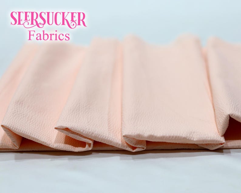 Puede incluir: Primer plano de tela seersucker melocot&oacute;n doblada. La tela tiene una superficie texturizada y est&aacute; dispuesta en un patr&oacute;n en capas. Las palabras "SEERSUCKER Fabrics" se muestran en rosa en la parte superior.
