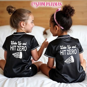 Pode incluir: Duas crian&ccedil;as vestindo pijamas de cetim preto com detalhes brancos e o texto "Wake Up and HIT ZERO" nas costas. Cada parte superior do pijama tem um gr&aacute;fico branco de um megafone com a palavra "CHEER" nele.