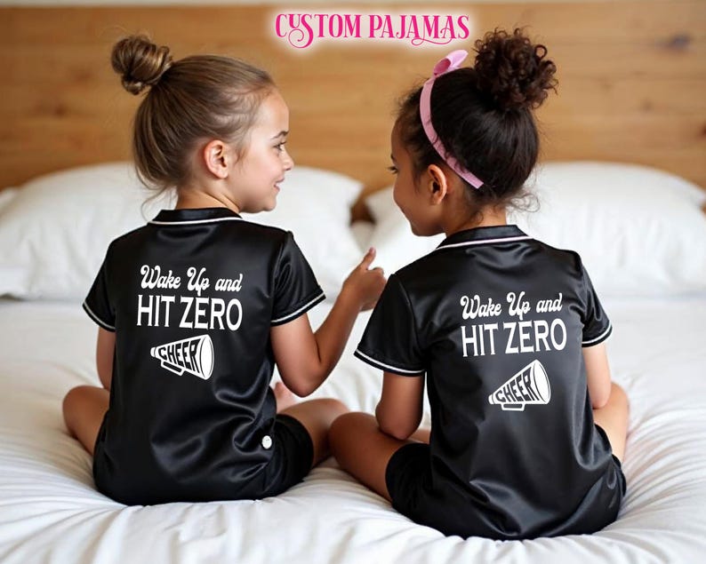 Pode incluir: Duas crian&ccedil;as vestindo pijamas de cetim preto com detalhes brancos e o texto "Wake Up and HIT ZERO" nas costas. Cada parte superior do pijama tem um gr&aacute;fico branco de um megafone com a palavra "CHEER" nele.