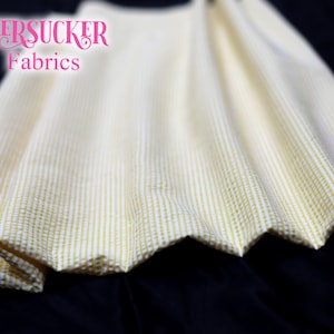 Puede incluir: Primer plano de tela seersucker con rayas verticales blancas y amarillo p&aacute;lido. La tela est&aacute; fruncida, mostrando un borde festoneado. El texto "SEERSUCKER Fabrics" es visible en rosa.
