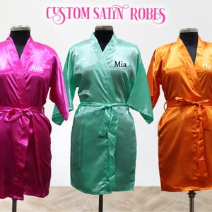 Könnte beinhalten: Drei Satin-Bademäntel in verschiedenen Farben: rosa, grün und orange. Jeder Bademantel hat einen Namen auf der Vorderseite gestickt: Ava, Mia und Dani.