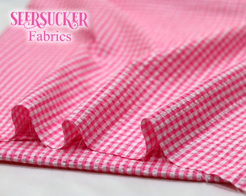 Puede incluir: Tela de seersucker rosa y blanca con un patr&oacute;n de cuadros. La tela est&aacute; doblada, mostrando la textura y el tejido. Las palabras "SEERSUCKER Fabrics" se muestran en una fuente rosa estilizada.