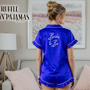 May include: Royal blue satin pajama set with white trim and the text "Bride to Be" on the back. The set includes a short-sleeved top and shorts. The words "Ruffle Satin Pajamas" are in the upper left corner.