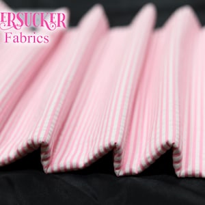 Puede incluir: Primer plano de tela seersucker a rayas rosas y blancas. La tela est&aacute; doblada en un patr&oacute;n ondulado, con el texto "SEERSUCKER Fabrics" en rosa en la parte superior izquierda. La tela probablemente se utiliza para ropa o decoraci&oacute;n del hogar.