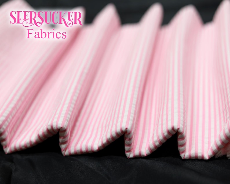 Puede incluir: Primer plano de tela seersucker a rayas rosas y blancas. La tela est&aacute; doblada en un patr&oacute;n ondulado, con el texto "SEERSUCKER Fabrics" en rosa en la parte superior izquierda. La tela probablemente se utiliza para ropa o decoraci&oacute;n del hogar.