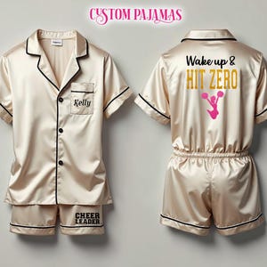 Pode incluir: Um pijama personalizado em cetim. A parte superior tem uma borda preta e um bolso com o nome "Kelly" bordado. A parte inferior tem as palavras "Cheer Leader" bordadas. A outra parte superior do pijama tem as palavras "Wake up & HIT ZERO" e um gr&aacute;fico de l&iacute;der de torcida rosa nas costas.