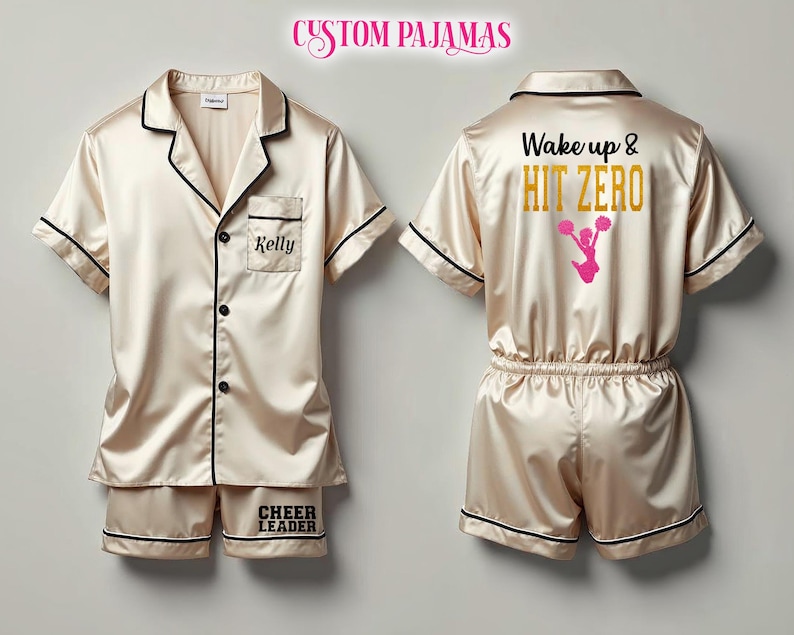 Pode incluir: Um pijama personalizado em cetim. A parte superior tem uma borda preta e um bolso com o nome "Kelly" bordado. A parte inferior tem as palavras "Cheer Leader" bordadas. A outra parte superior do pijama tem as palavras "Wake up & HIT ZERO" e um gr&aacute;fico de l&iacute;der de torcida rosa nas costas.