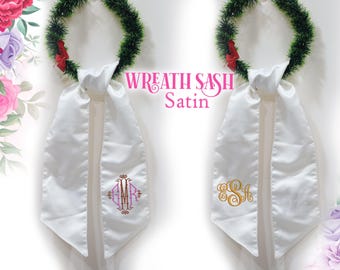 Könnte beinhalten: Zwei weiße Satin-Kranzschärpen mit gestickten Monogrammen. Jede Schärpe ist an einem grünen Kranz mit einer roten Schleife befestigt. Der Text "Wreath Sash Satin" ist in Pink dargestellt. Eine Schärpe hat ein rosa-goldenes Monogramm, die andere ein goldenes Monogramm.