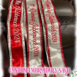 May include: Three custom embroidered sashes. One is red with "Miss Jabberwock 2024" in red script. The second is white with "1st Runner up Jabberwock 2024" in red script. The third is white with "2nd Runner up Jabberwock 2024" in red script. The text "CUSTOM EMBROIDERY SASH" is at the bottom.